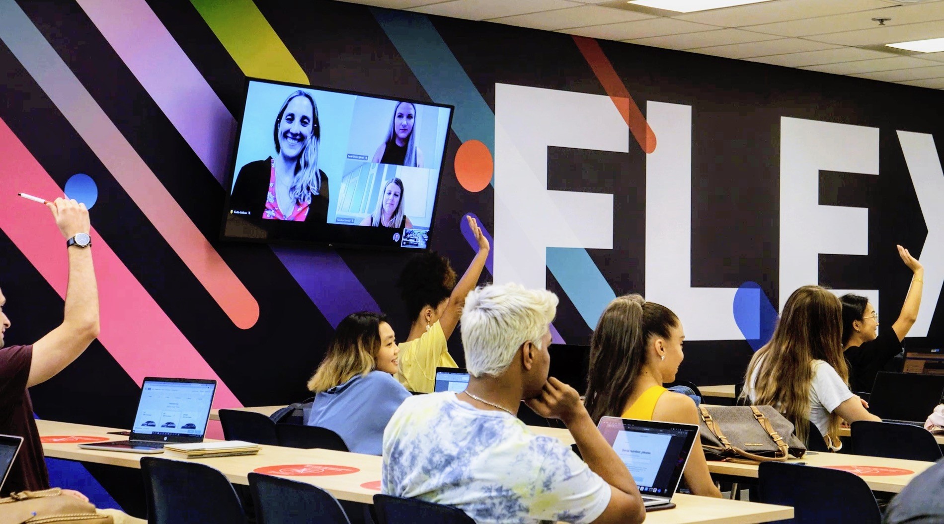 Des étudiants dans une classe Flex. Le mur du côté de la salle est ornée d’une murale colorée où on lit « Flex ». Un grand écran fixé au mur permet de voir des personnes qui participent à distance. Dans la classe, plusieurs étudiantes ont la main levée. La plupart des élèves présents ont un ordinateur portable.