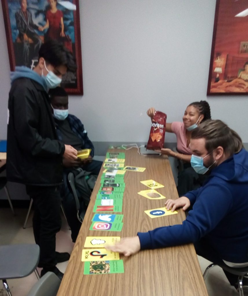 Une étudiante montre fièrement à la caméra un sac de Doritos. 3 étudiants masqués autour de la même table qu'elle manipule des cartes jaunes et vertes.