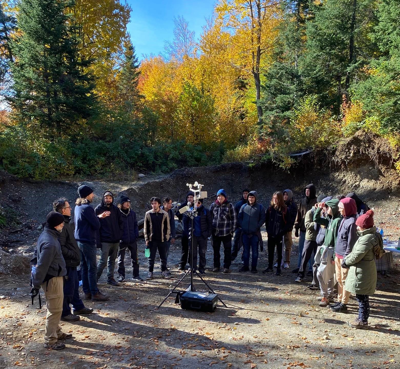  About 20 people are standing in a semicircle around a portable weather station on a tripod in the center of a sandy clearing surrounded by fall-coloured hardwoods. The people are listening to a man, whom we guess is a teacher explaining something to them.