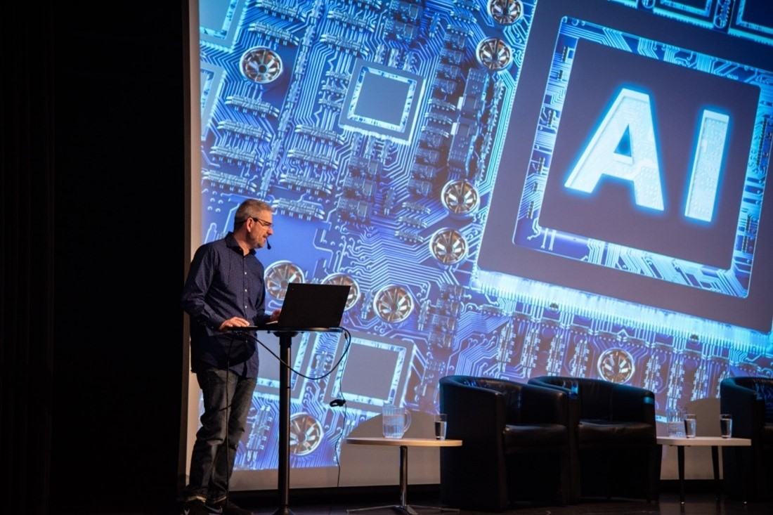 Édouard Morrissette devant un lutrin. Un écran de projection derrière lui montre une image des lettre « AI » au centre d’un circuit imprimé