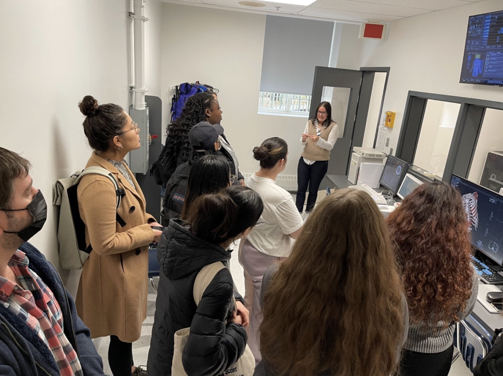 Facing away from the camera, 9 college students are standing in a medical imaging technology laboratory equipped with several computer screens on which scan results are displayed. They are listening to their female teacher, who is giving explanations.
