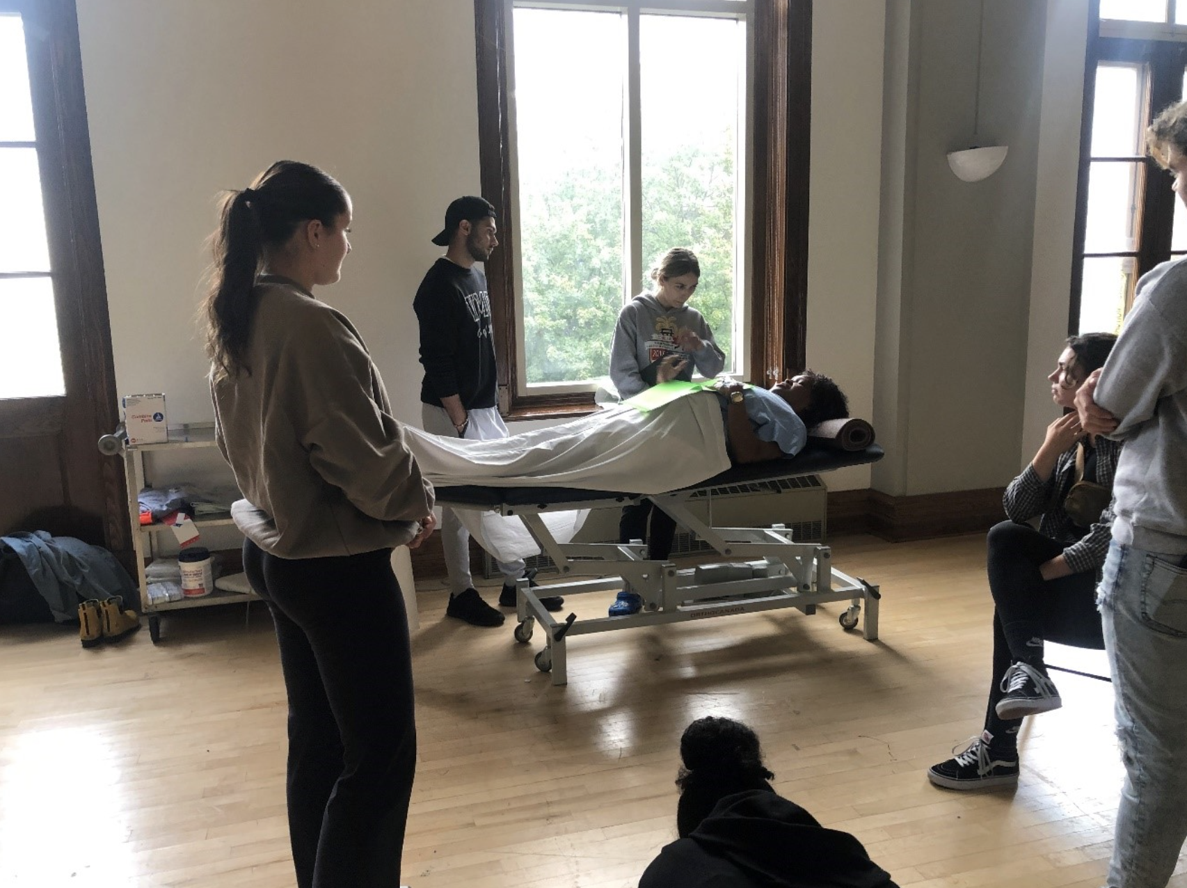 In a classroom with big windows, a female college student is standing beside a stretcher. On the stretcher is another female, dressed in a hospital gown and role-playing a patient. The student appears to be listening or talking to the patient. 5 college students, one of which is sitting down while the others are standing up, are observing the scene.
