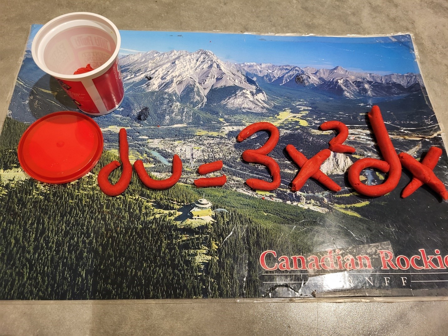 Un pot de pâte à modeler rouge est ouvert et posé sur un napperon vintage montrant une photo des rocheuses, sur lequel on lit "Canadian Rockies. Banff". De la pâte à modeler a été utilisée pour former les symboles d u = 3 x exposant 2 d x.