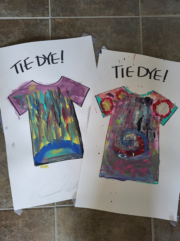 2 cartons sont posés sont un plancher en céramique. Sur chaque carton, quelqu'un a peint les mots «Tie Dye!» et la forme d'un t-shirt est dessinée en noir. L'intérieur des formes de chandails sont peintes d'une multitude de couleurs, imitant le style tie-dye.