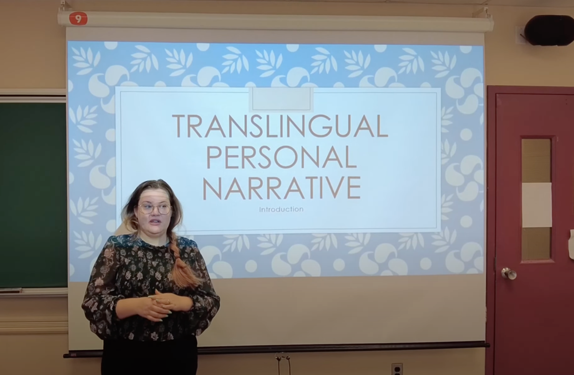 April Passi is standing up in front of a classroom. Behind her, we can see a pull-down projector screen displaying the words “Translingual personal narrative.” On the left-hand side, we can see a blackboard, while on the right, we can see a door.
