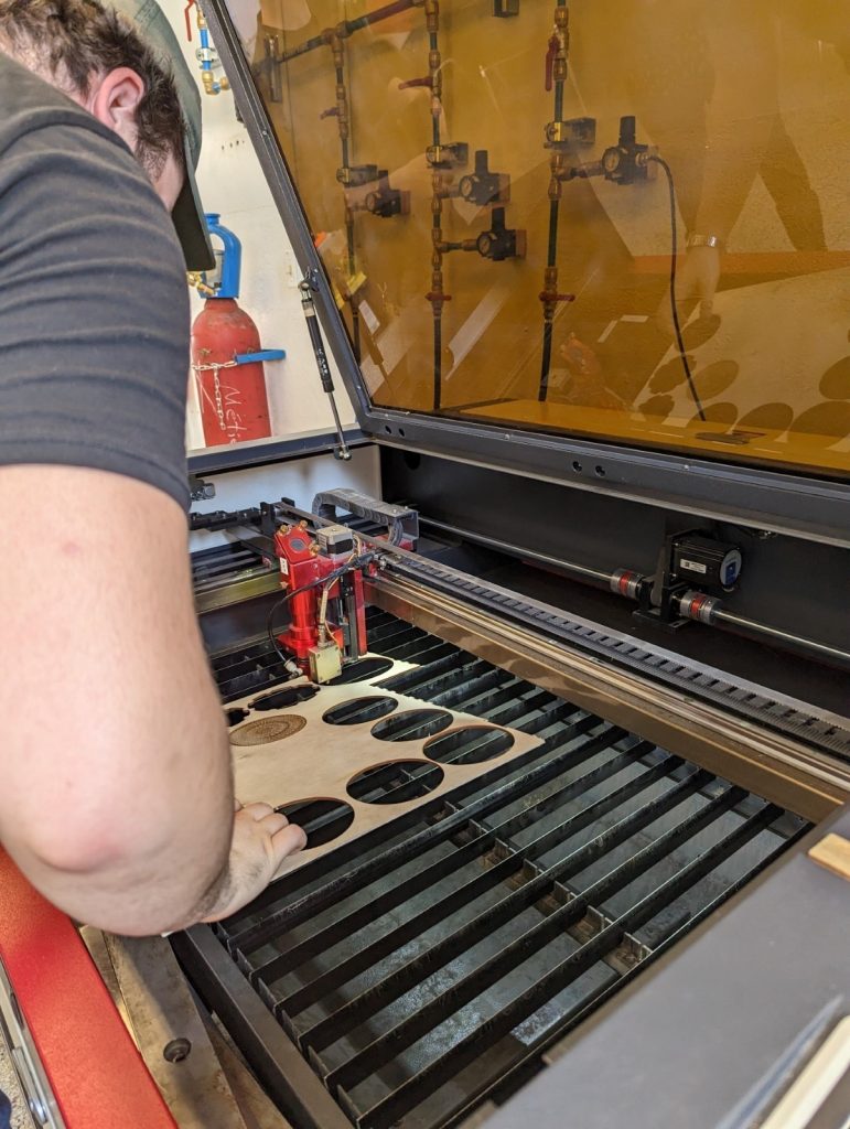Close-up photo shot showing a metal grid on the inside of a laser engraver cutting machine of which the protective cover is open. We see the right arm and the back of the head of a male student who is placing a thin piece of wood onto the grid, aligning it with the machine’s laser. Image source: Andy Van Drom