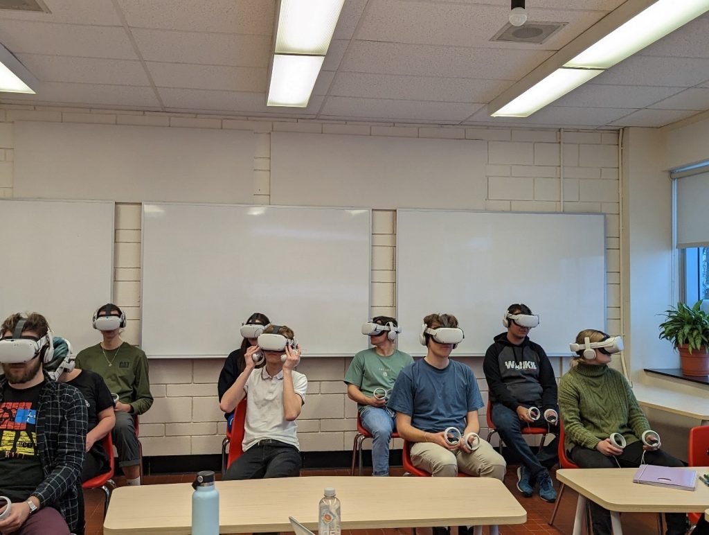 In the back of a classroom, in front of a wall covered in 3 whiteboards, 9 students are sitting on chairs that are lined up in 2 rows, with about 5 students in each row. All students are wearing virtual reality headsets. Image source: Andy Van Drom