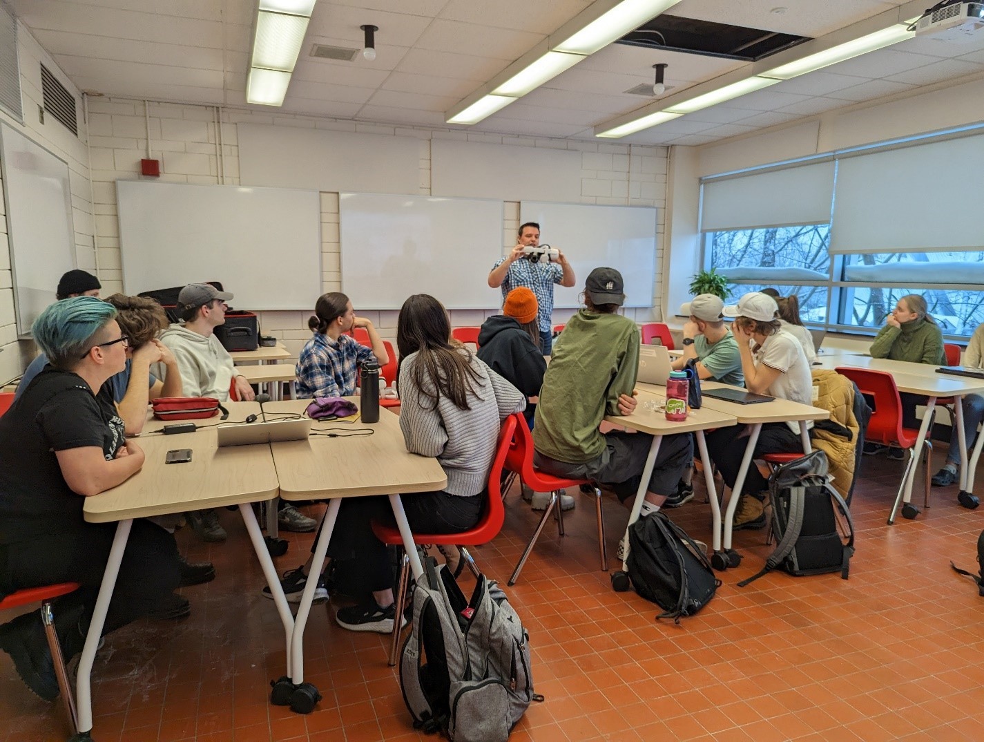 Photo of a classroom with whiteboards covering the left and back walls, and windows on the right wall. Students are seated around 3 islands of 5 or 6 individuals. In the back of the room, a male technician is holding up a virtual reality headset while talking to the students. It can be inferred that he is explaining to the students how to use the headset. Image source: Andy Van Drom