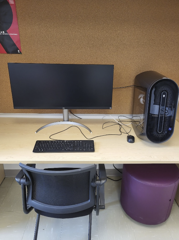 Un ordinateur de bureau est posé sur un bureau. Une chaise de travail est placée devant le bureau, et un tabouret est glissé sous le bureau. Un babillard est fixé au mur derrière le bureau.