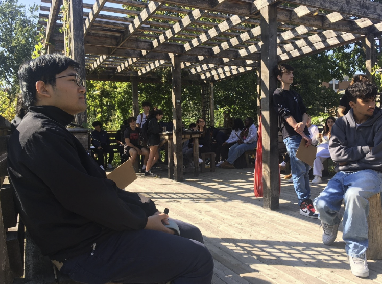 Young adult college students are standing up and sitting on benches under a wooden pergola. Some of them are holding clipboards and pens. Their facial expressions suggest they are listening to someone, who is outside the frame of the image.
