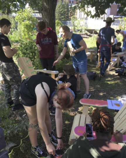 Young adult college students are standing up and bending over several Adirondack chairs dispersed in a wooded area. They are holding paint brushes and referring to plans and their cell phone to paint the natural wooden chairs in bright colours and patterns.